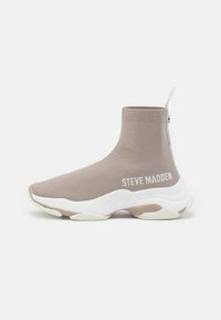 Zapatilla de punto beige con diseño de tobillo alto, suela blanca y logo "STEVE MADDEN". Presenta una superficie texturizada y una lengüeta en la parte trasera.