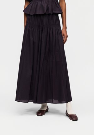 Frau trägt einen langen schwarzen plissierten Rock, weiße Strümpfe und dunkelbraune flache Schuhe mit quadratischer Spitze, steht vor einem einfarbigen Hintergrund.