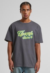 Grå oversized t-shirt lavet af bomuld med "Upscale Studios" grafik i lys gul og grøn, med en afslappet pasform og korte ærmer.