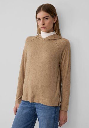 Helle braune Langarm-Hoodie mit V-Ausschnitt und seitlichen Schlitzen, aus weichem Material. Kombiniert mit blauen Jeans im lässigen Schnitt.
