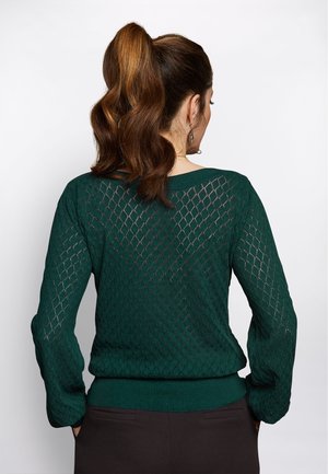 Femme avec une queue de cheval brune ondulée portant un pull en maille léger vert foncé et un pantalon sombre, debout les mains dans les poches, tournée de dos.