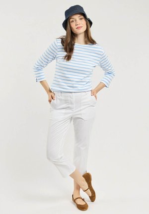 Femme portant une chemise à rayures bleues à manches longues, un pantalon blanc court, des chaussures marron et un chapeau bob bleu foncé, debout les mains dans les poches.