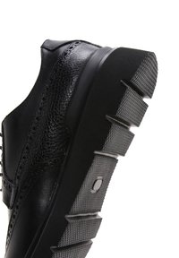 Chaussure en cuir noir avec des accents texturés et une épaisse semelle en caoutchouc gris, dotée d'un profil de semelle à motifs et de détails de brogue élégants.