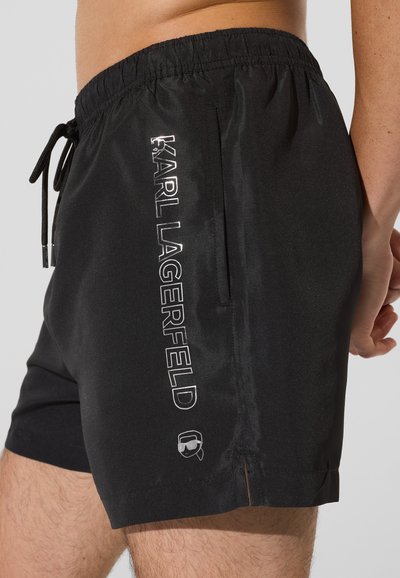 Short de bain noir en matière légère, avec une taille élastique, un cordon de serrage et un logo réfléchissant "KARL LAGERFELD" sur le côté.
