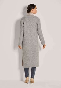 Frau in einem langen grauen Sweatermantel, dunklen Jeans und beigen High Heels, die vor einem einfarbigen hellen Hintergrund steht und von hinten schaut.