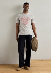 Gray short-sleeve t-shirt with red circular logo, paired with dark blue straight-leg pants and beige footwear, holding a brown jacket.