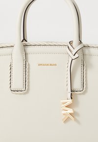 Weiße Lederhandtasche mit geflochtenen Griffen, goldfarbener Logo-Beschriftung und charmantem Anhänger. Verfügt über ein strukturiertes Design und eine glatte Textur.