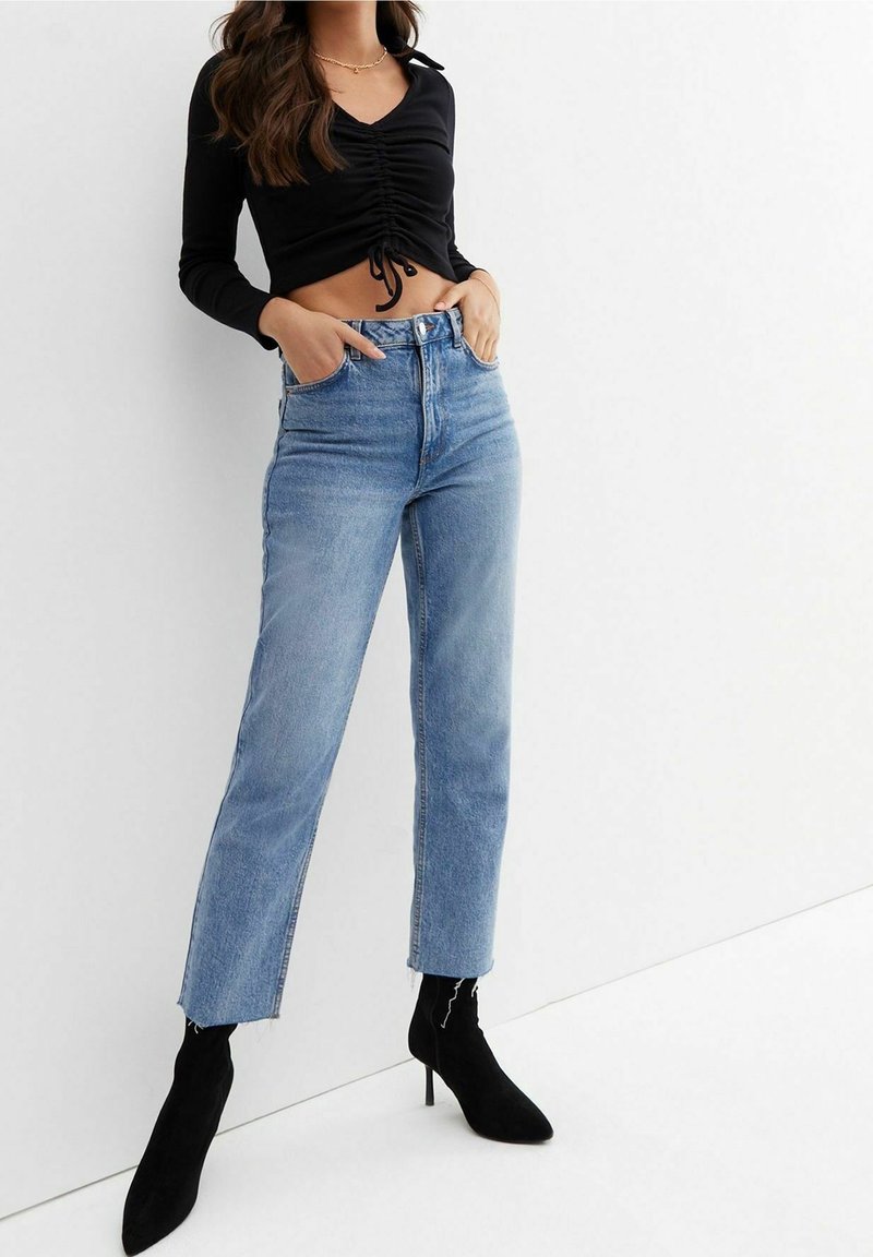 Schwarzes langärmeliges, cropped Oberteil mit gerafften Details vorne, kombiniert mit hochtaillierten, hellblauen, geraden Jeans und schwarzen Knöchel Stiefeln.
