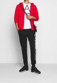 Sudadera con capucha roja con cremallera sobre una camiseta blanca con un diseño gráfico, combinada con pantalones de chándal negros con logotipos blancos y zapatillas negras.