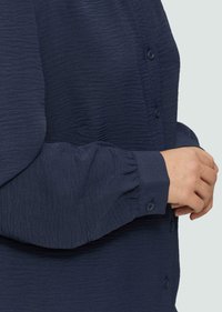 Marineblaue Hemdbluse aus strukturiertem Stoff, mit langen Ärmeln und gerafften Manschetten, sowie einer Knopfleiste vorne.