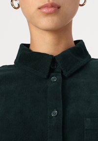 Camicia di velluto a coste verde scuro con colletto a punta, chiusura a bottoni e tasca frontale con texture a coste. Luce che si riflette sul tessuto.