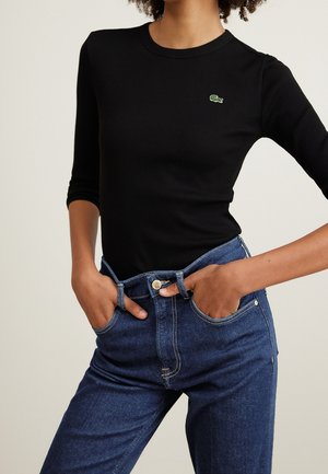 Vrouw draagt een aangesloten zwart geribd shirt met lange mouwen en een klein groen krokodillenlogo, ingestopt in een high-waisted blauwe spijkerbroek, met handen in de zakken.