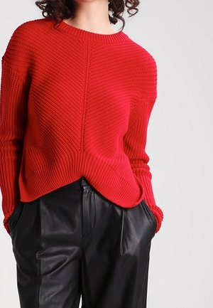 Mujer con un suéter de punto texturizado rojo con dobladillo puntiagudo y pantalones de cuero negros de cintura alta, con las manos en los bolsillos, sobre un fondo claro.