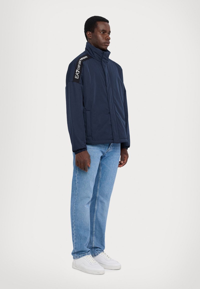Chaqueta ligera azul marino con cuello alto, frontal con cremallera y puños elásticos. Presenta detalles de logo en los hombros y bolsillos laterales.