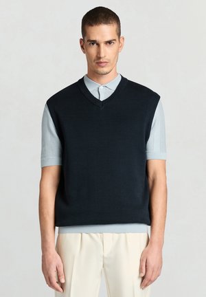 Gilet en tricot bleu marine avec un décolleté en V, des manches courtes et des bords côtelés, porté sur une chemise à col bleu clair en tissu texturé.