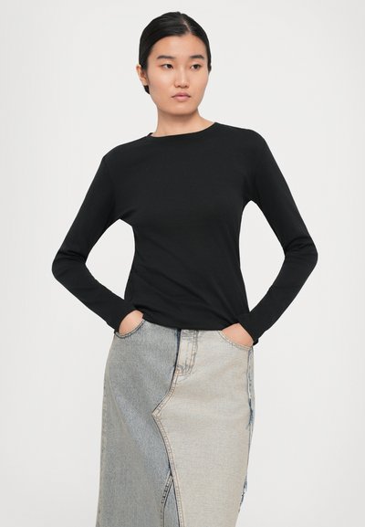 Kvinna i svart långärmad tröja och blå-beige lapptäckad jeanskjol står med händerna i fickorna och tittar åt sidan.
