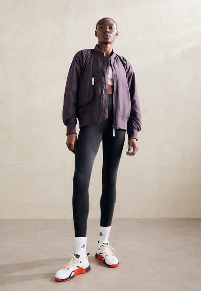 Lila bomberdzseki cipzáras zsebekkel, fekete leggings és fehér/piros sportcipő. Simább anyag, bordázott mandzsetták és rövidített fazon.