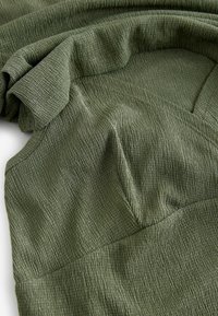 Grüner texturierter Stoff mit einer zerknitterten Erscheinung, der drapierte Falten und eine glatte Oberfläche zeigt. Nahtdetails sind an der Außenseite sichtbar.