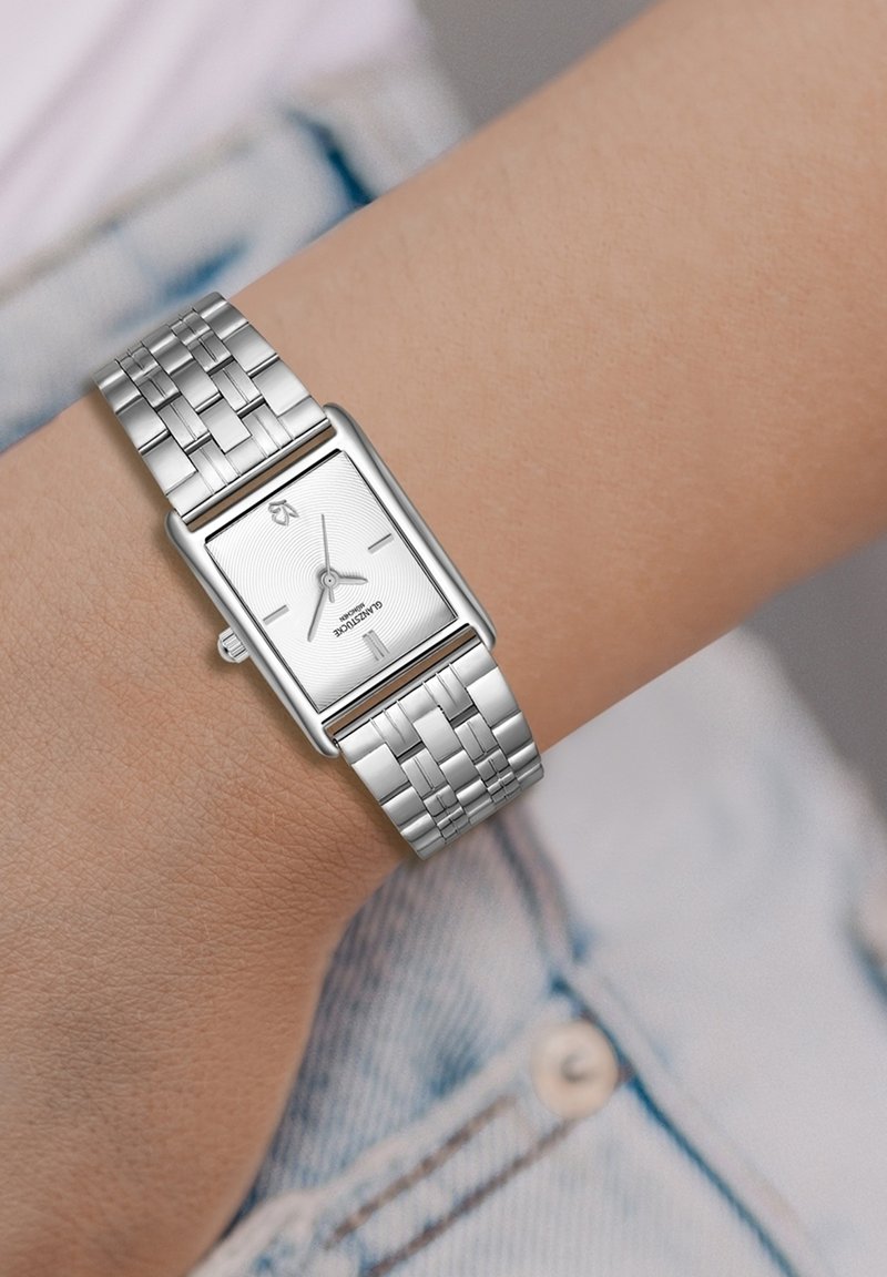 Hõbedane ristkülikukujuline kell tekstureeritud valge näidikuga, metallist link-ranneke ja minimalistlik disain. Kantud kerge nahatooni randmel.