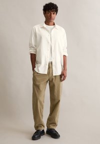Biała koszula na guziki, nałożona na jasną koszulę, zestawiona z jasnobrązowymi spodniami sztruksowymi o prążkowanej fakturze i czarnymi masywnymi butami. Neutralne tło.