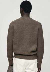 Brauner Strickpullover mit Rundhalsausschnitt, der eine strukturierte Oberfläche und gerippte Bündchen aufweist. Kombiniert mit hellen Hosen.
