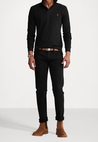 Czarna koszulka polo z długim rękawem i kołnierzykiem, zestawiona z czarnymi jeansami slim-fit i brązowymi zamszowymi botkami za kostkę. Brązowy pasek z silikonową klamrą.