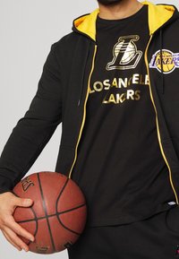 Svart zip-up hoodie med gula accenter, kombinerad med en svart T-shirt som har guldtext och logotyp för "Los Angeles Lakers"; håller en basketboll.