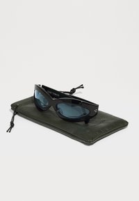 Lunettes de soleil noires avec des verres bleus, au design élégant, dotées de petites touches en métal, reposant sur un étui vert texturé avec un système de fermeture à cordon.