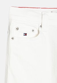 Valkoiset farkut, joissa on sivutasku, jossa on pieni brodeerattu logo punaisella, valkoisella ja sinisellä, sekä metalliset napin yksityiskohdat.