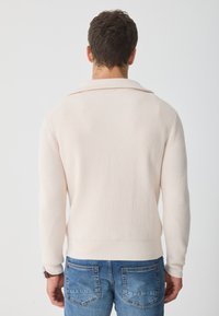 Világos bézs kötött pulóver bordázott struktúrával, széles gallérral és szűkített mandzsettával, kék farmernadrággal párosítva.