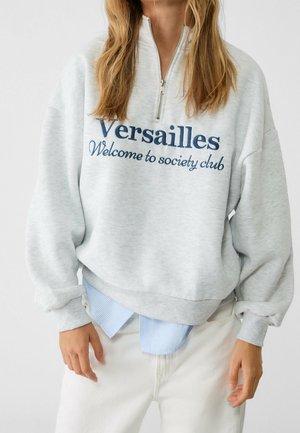 Persoon draagt een lichtgrijze half-ritssluiting trui met de tekst "Versailles Welcome to society club", laagjes over een blauw overhemd en witte broek.