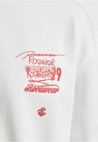 Bílá mikina se žlutými logy a textem. Obsahuje mix písem s výrazně zobrazeným nápisem "Rocawear". Bavlněná textura.