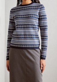 Pull bleu marine à motifs avec manches longues et col rond, présentant des motifs géométriques blancs. Associé à une jupe grise.