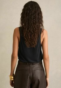 Donna con lunghi capelli ricci scuri indossa un top nero senza maniche, pantaloni di pelle marroni e diversi braccialetti dorati, rivolta verso una parete beige chiaro.