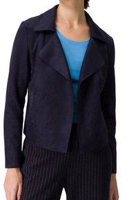 Nainen, joka käyttää tummansinistä avointa mokkajakku yli vaaleansinisestä paidasta ja mustista raidallisista housuista valkoista taustaa vasten.