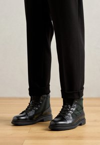 Juodi odiniai ir audinių raištelių batai, dėvimi su apvertais juodais kelnių, stovint ant medinio grindų.