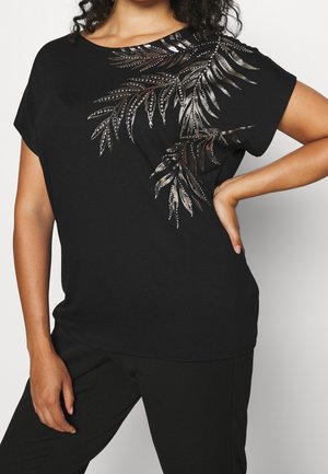 Kobieta ubrana w czarną bluzkę z krótkim rękawem, ozdobioną metalicznymi wzorami liści na ramieniu i górnej części klatki piersiowej, w połączeniu z czarnymi spodniami.