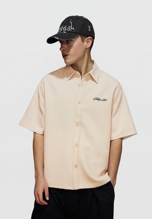 Fiatal férfi, bézs színű, rövid ujjú, hímzett gombos ingben és fekete, kopott baseballsapkában, oldalra nézve.