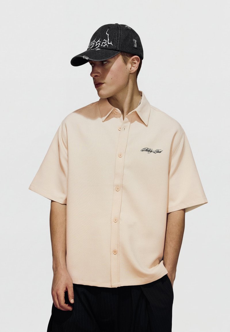 Jeune homme portant une chemise beige à manches courtes avec des broderies et une casquette de baseball noire usée, regardant sur le côté.