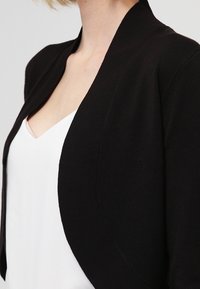 Černý pletený kardigan s otevřeným předním dílem, jemnou texturou a zakončením s obloukovým okrajem, nošený přes bílý top.