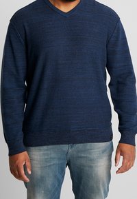 Navy knitted sweater with a textured pattern, V-neck design, ribbed cuffs, and hem; paired with light blue jeans.