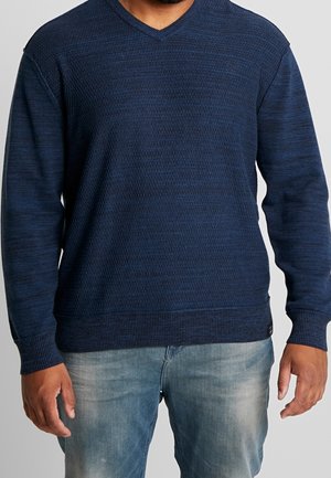 Maglione lavorato a maglia blu navy con un motivo texture, design a V, polsini e orlo a coste; abbinato a jeans azzurri chiari.