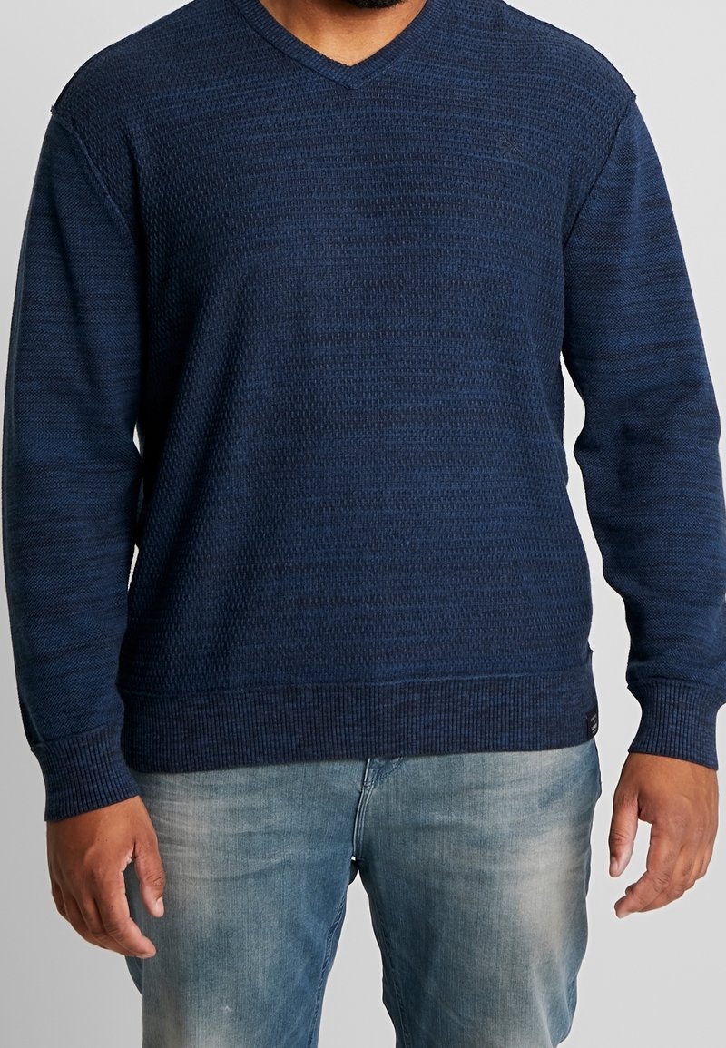 Navy knitted sweater with a textured pattern, V-neck design, ribbed cuffs, and hem; paired with light blue jeans.
