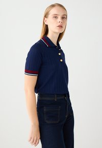 Polo shirt navy con texture a coste, maniche corte e bottoni dorati. Il colletto presenta dettagli rossi e bianchi. Abbinato a jeans di denim scuro.