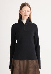 Maglione nero a costine con colletto a mezze zip e maniche lunghe, abbinato a una gonna texturizzata marrone, su uno sfondo semplice.