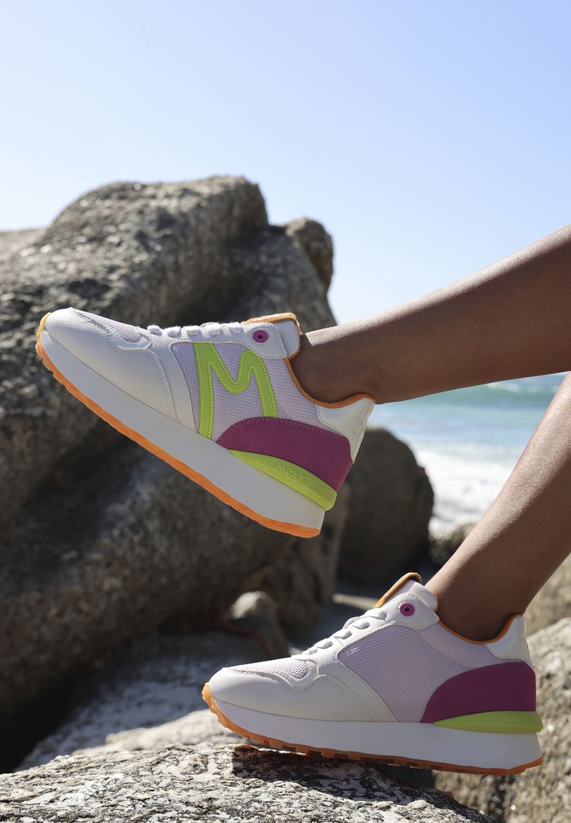 Witte sneakers van mesh en synthetische materialen, met groene en paarse accenten, een oranje zool en een opvallend 'M'-logo aan de zijkant.