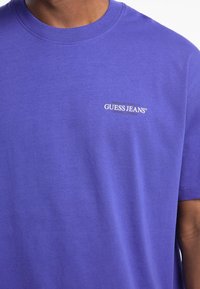 Vijolična bombažna majica s krožnim izrezom in kratkimi rokavi. Na sprednji strani se nahaja bel napis "GUESS JEANS". Gladka tekstura, brez vzorcev.