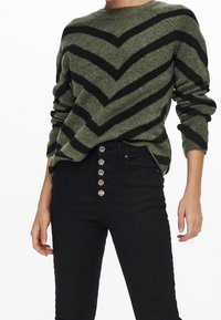 Pull vert olive avec des rayures en chevron noires, texture tricotée et col rond. Associé à un jean noir taille haute avec des accents de boutons.