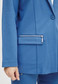Modrá sako s elegantním designem, hladkým materiálem, přední kapsou na zip a knoflíkovým zapínáním. Rukávy končí jednoduchým manžetou.