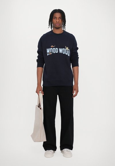 Hanorac bleumarin cu text "WOOD WOOD" și broderie cu fluture; asortat cu pantaloni negri și o geantă de umăr de o nuanță mai deschisă. Încălțăminte casual vizibilă.